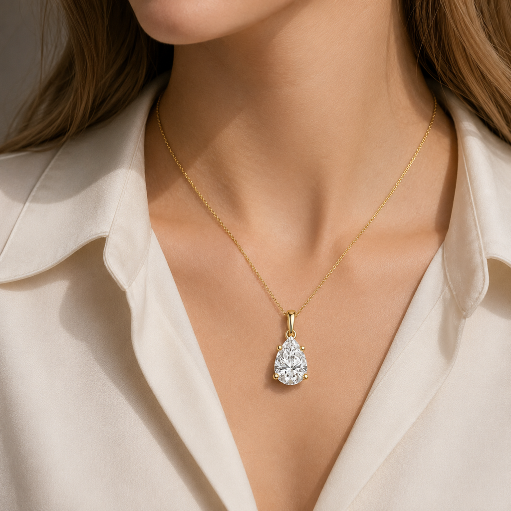 A close-up of a model wearing a delicate gold chain necklace with a pear-cut clear gemstone pendant, styled with a cream-colored collared shirt.
