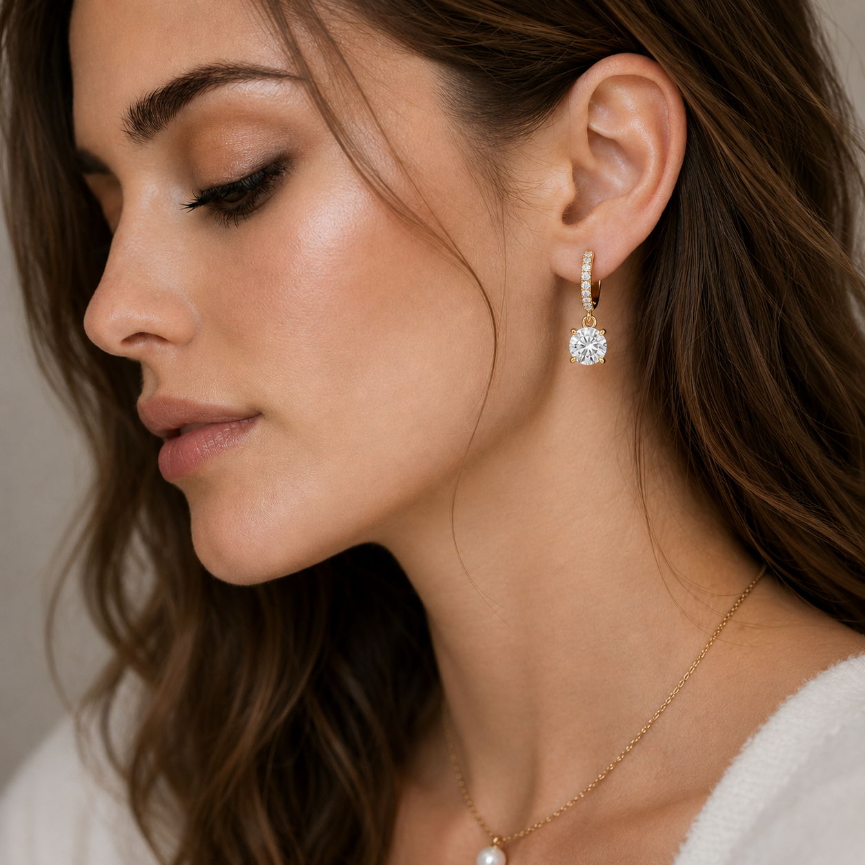 A close-up side profile of a woman wearing a delicate gold drop earring with a clear round gemstone and a thin pavé-set top, paired with a fine gold chain necklace with a pearl. The background is a soft, neutral beige.