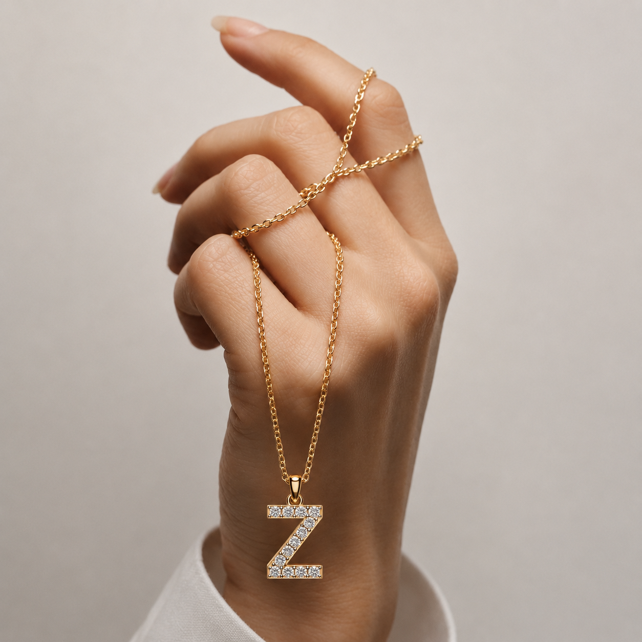 A gold chain necklace with a letter 'Z' pendant paved with small clear stones, shown worn on a hand against a soft light gray background.