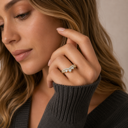 A close-up lifestyle shot of a woman wearing a delicate gold band ring set with small clear stones, including a central round stone. The image focuses on her hand and the ring, with a soft beige background.