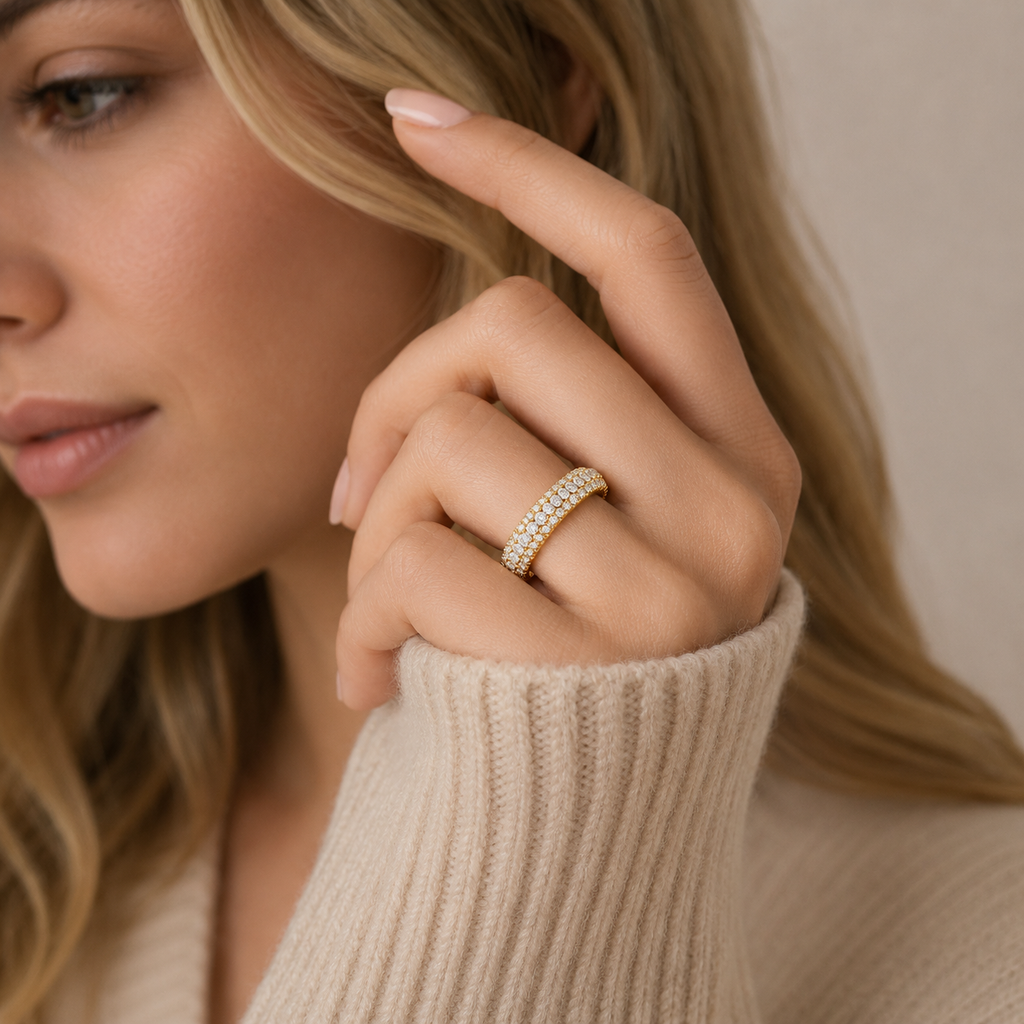 A close-up lifestyle shot of a woman wearing a delicate gold ring set with small clear stones on her ring finger. She is wearing a beige ribbed knit sweater, and the background is a soft, neutral beige wall.