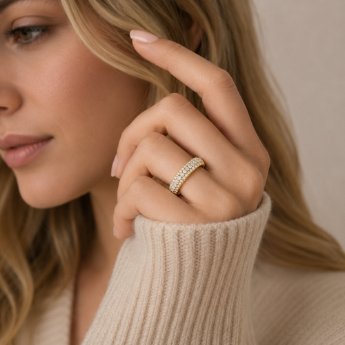 A close-up lifestyle shot of a woman wearing a delicate gold ring set with small clear stones on her ring finger. She is wearing a beige ribbed knit sweater, and the background is a soft, neutral beige wall.