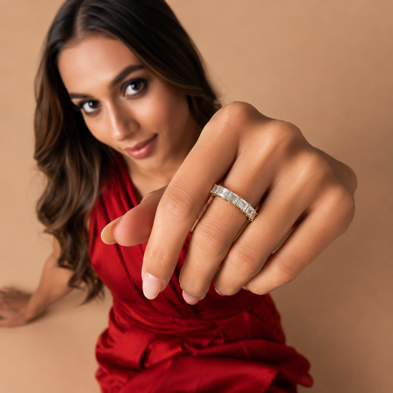 A woman in a red dress extends her hand toward the camera, wearing a silver band ring with rectangular clear stones on her ring finger, set against a warm beige background.
