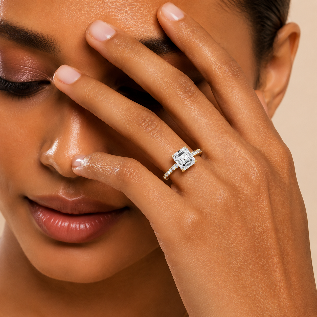 A close-up lifestyle shot of a model wearing a silver engagement ring with a rectangular clear gemstone on her finger, with her hand resting near her face against a warm beige background.