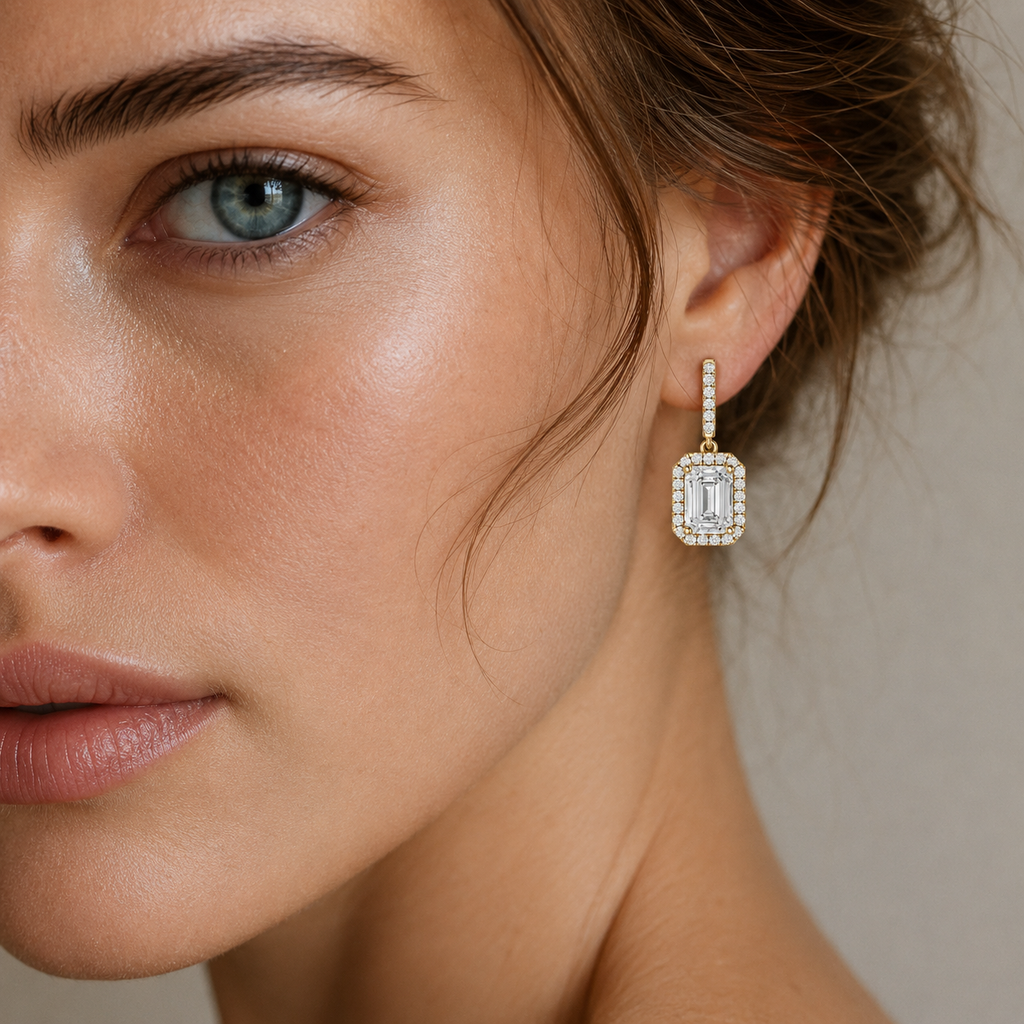 A close-up of a woman's face and ear wearing a single rectangular drop earring with a halo of small clear stones and a larger central clear stone, photographed against a soft neutral background.