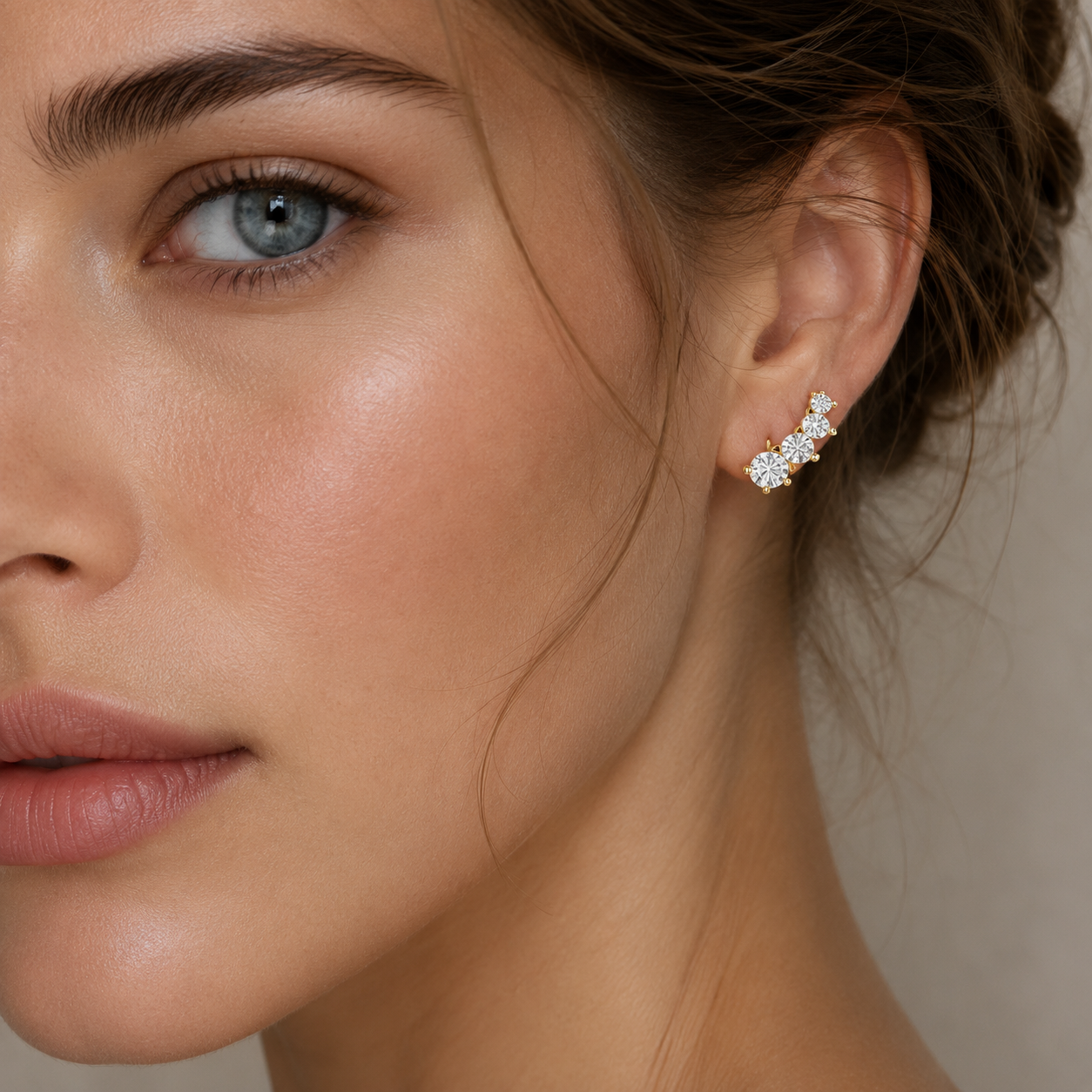 A close-up of a woman's ear wearing a small cluster stud earring with clear stones and a gold-toned setting against a soft beige background.