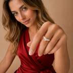 A woman in a red satin dress extends her hand to show a delicate silver ring with a small cluster of clear gemstones on her ring finger, set against a warm beige background.