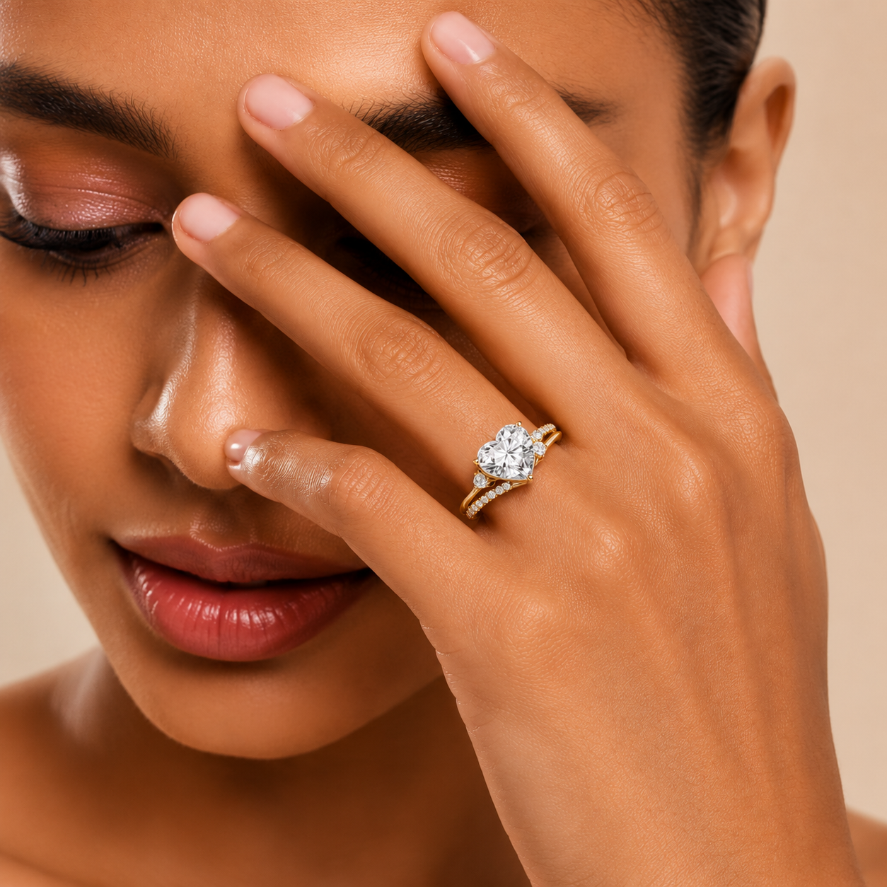 A close-up lifestyle shot of a model wearing a gold ring with a heart-shaped clear gemstone on her finger, with her hand resting against her face.