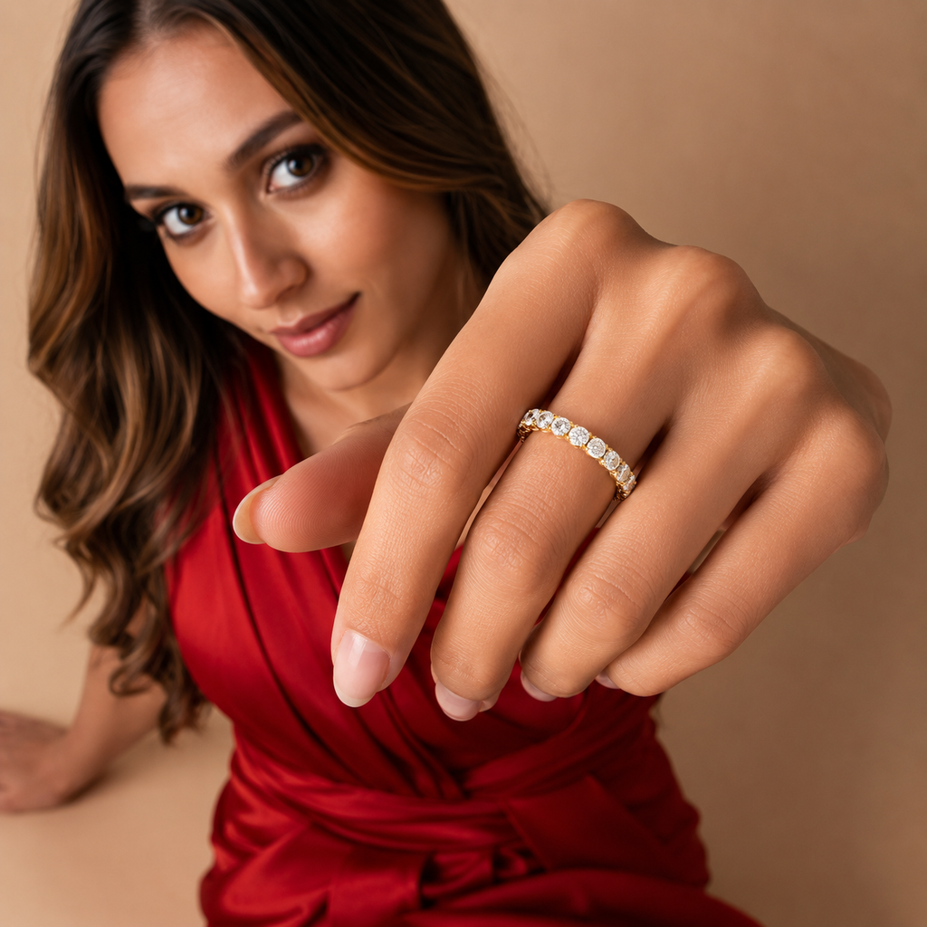 A close-up lifestyle shot of a woman wearing a delicate gold band ring with small clear stones, pointing toward the camera. She is wearing a red dress against a solid beige background.