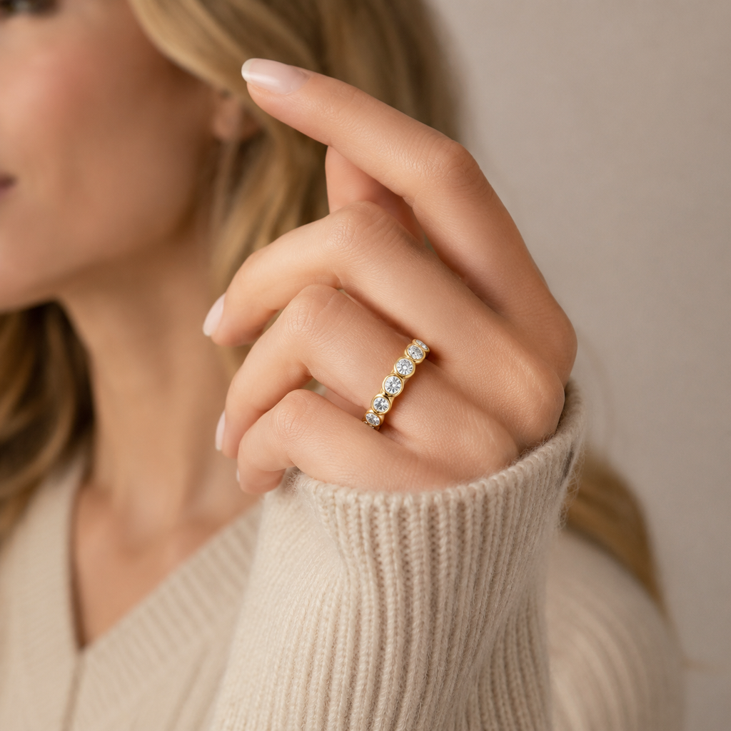 A close-up of a woman's hand wearing a delicate gold band ring set with small clear stones, styled with a beige ribbed knit sweater against a soft neutral background.