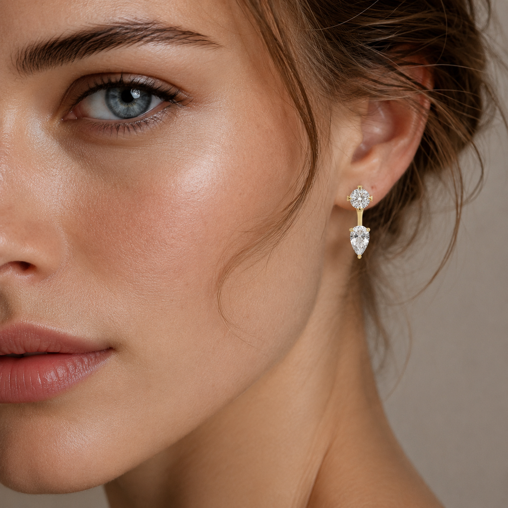 A close-up of a woman's face and ear wearing a gold drop earring with a clear teardrop-shaped gemstone.