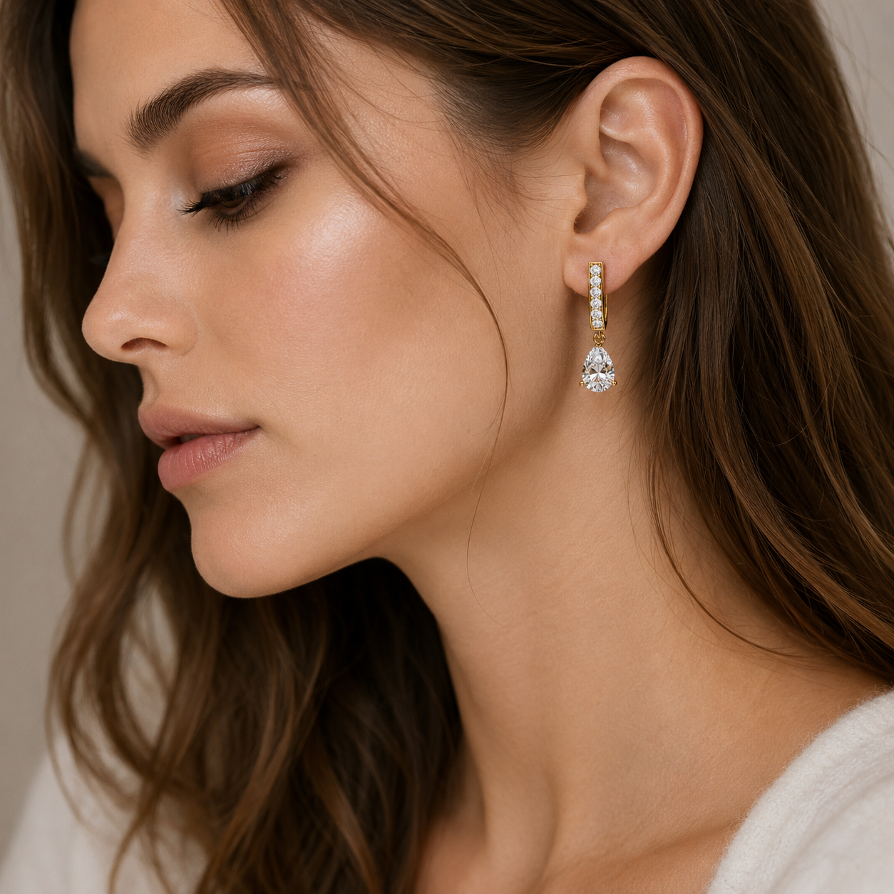 A close-up side profile of a woman wearing a delicate gold drop earring with a small vertical row of clear stones and a teardrop-shaped clear gemstone dangling at the end.