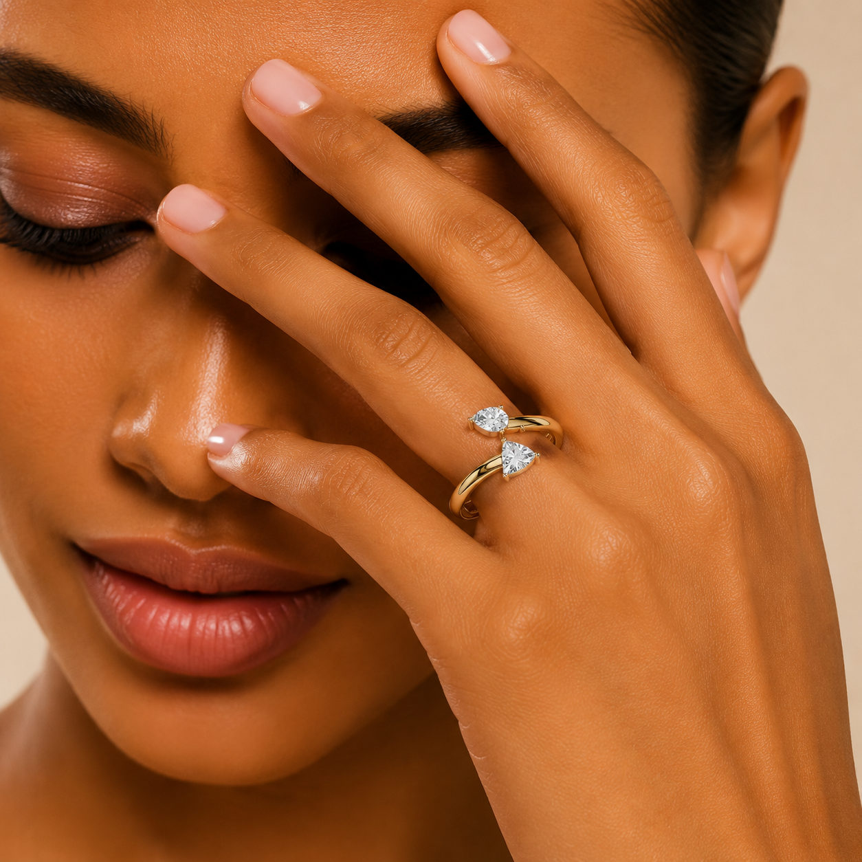 A close-up lifestyle shot of a model's hand wearing a gold ring with two clear heart-shaped gemstones, set against a warm beige background.