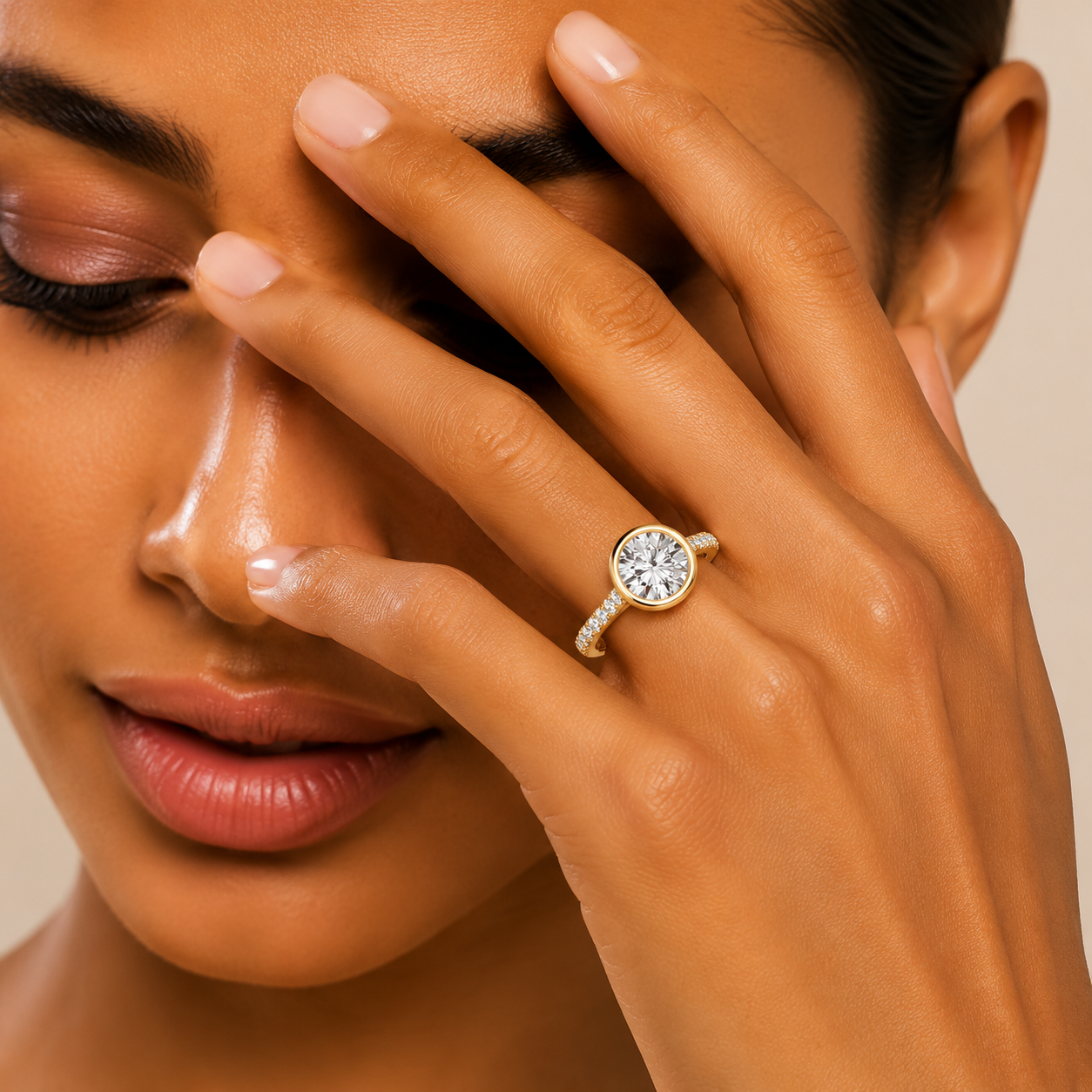 A close-up lifestyle shot of a model wearing a gold engagement ring with a round-cut clear gemstone on her finger, with her hand resting against her face.