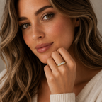 A close-up lifestyle shot of a woman with wavy brown hair wearing a gold band ring set with small clear stones on her ring finger. She is wearing a cream-colored knit sweater and looking directly at the camera.