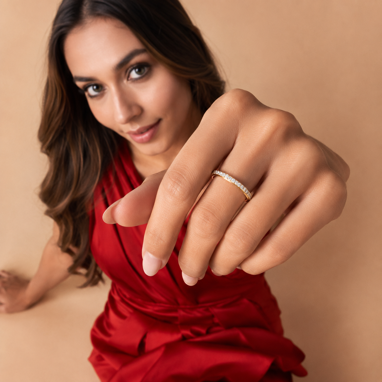A woman in a red dress extends her hand toward the camera, wearing a thin gold band ring set with small clear stones on one finger, against a warm beige background.