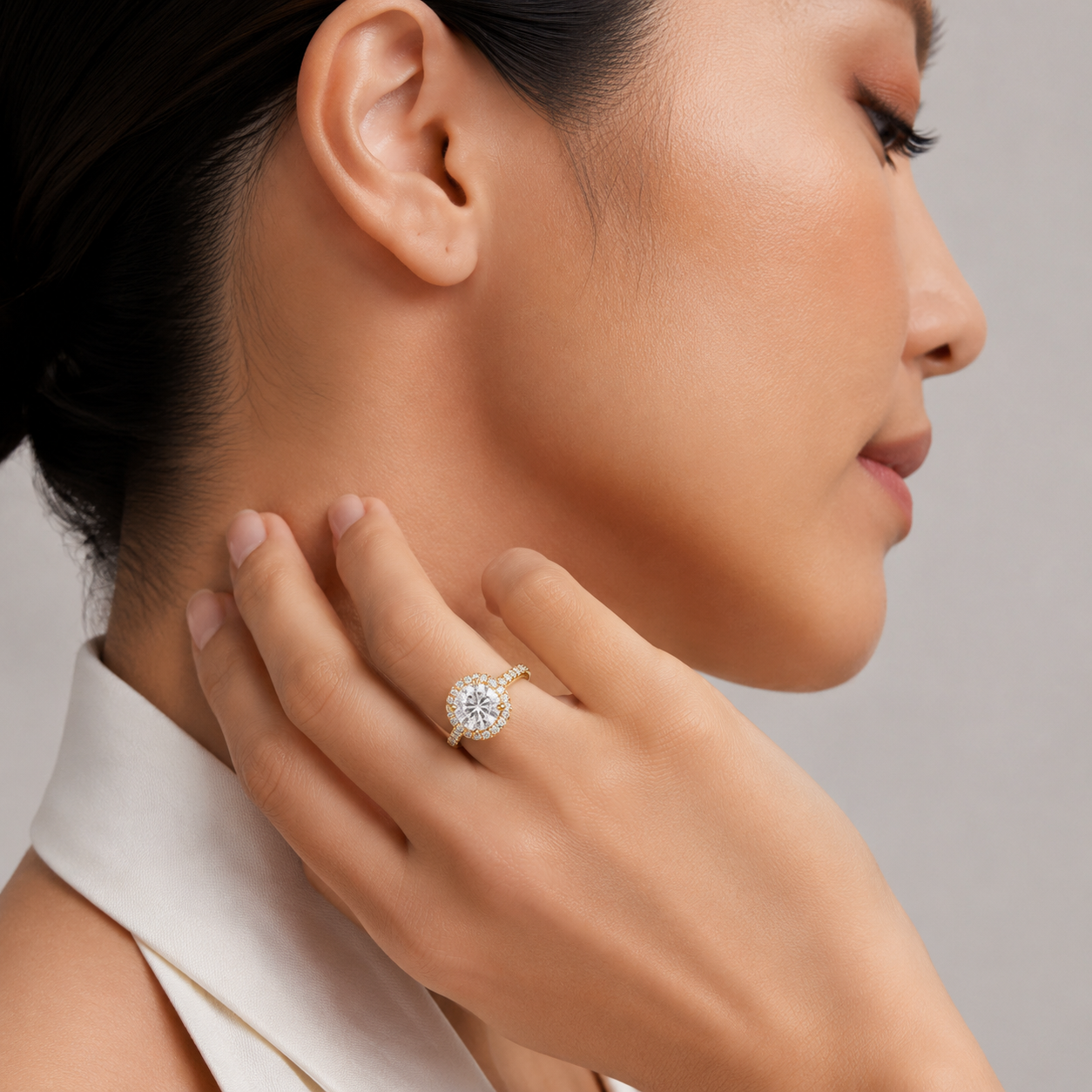 A close-up lifestyle shot of a woman wearing a yellow gold halo ring with a round-cut clear center stone and surrounding pavé setting. The ring is shown on her finger as she gently touches her neck, wearing a white collar against a neutral gray background.