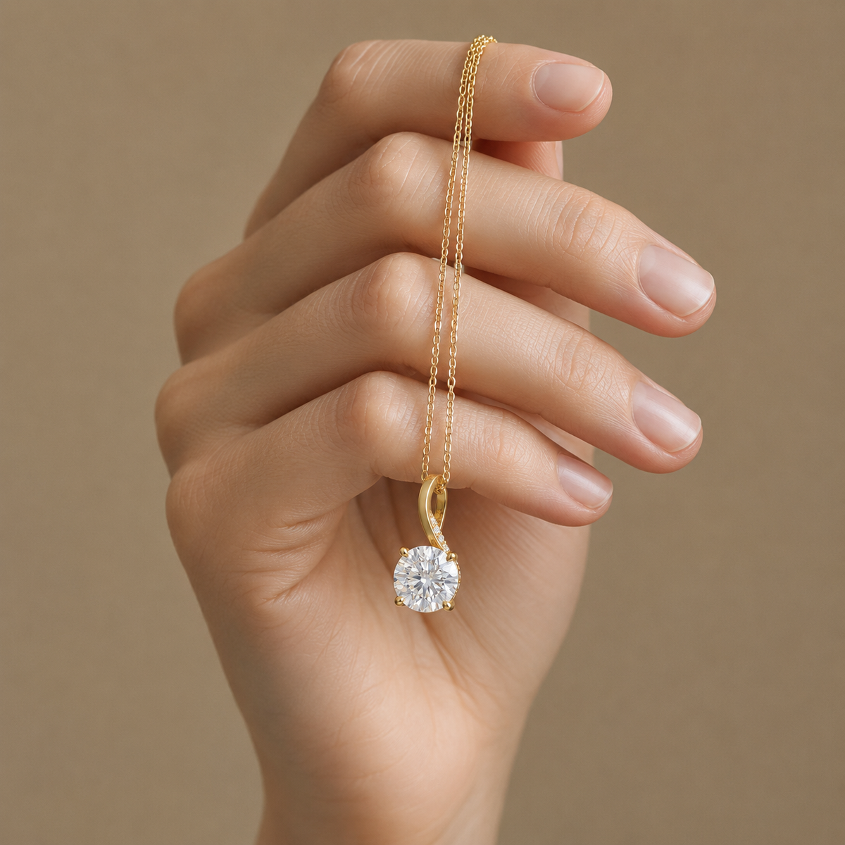 A close-up of a hand holding a delicate gold chain necklace with a round-cut clear gemstone pendant against a soft beige background.