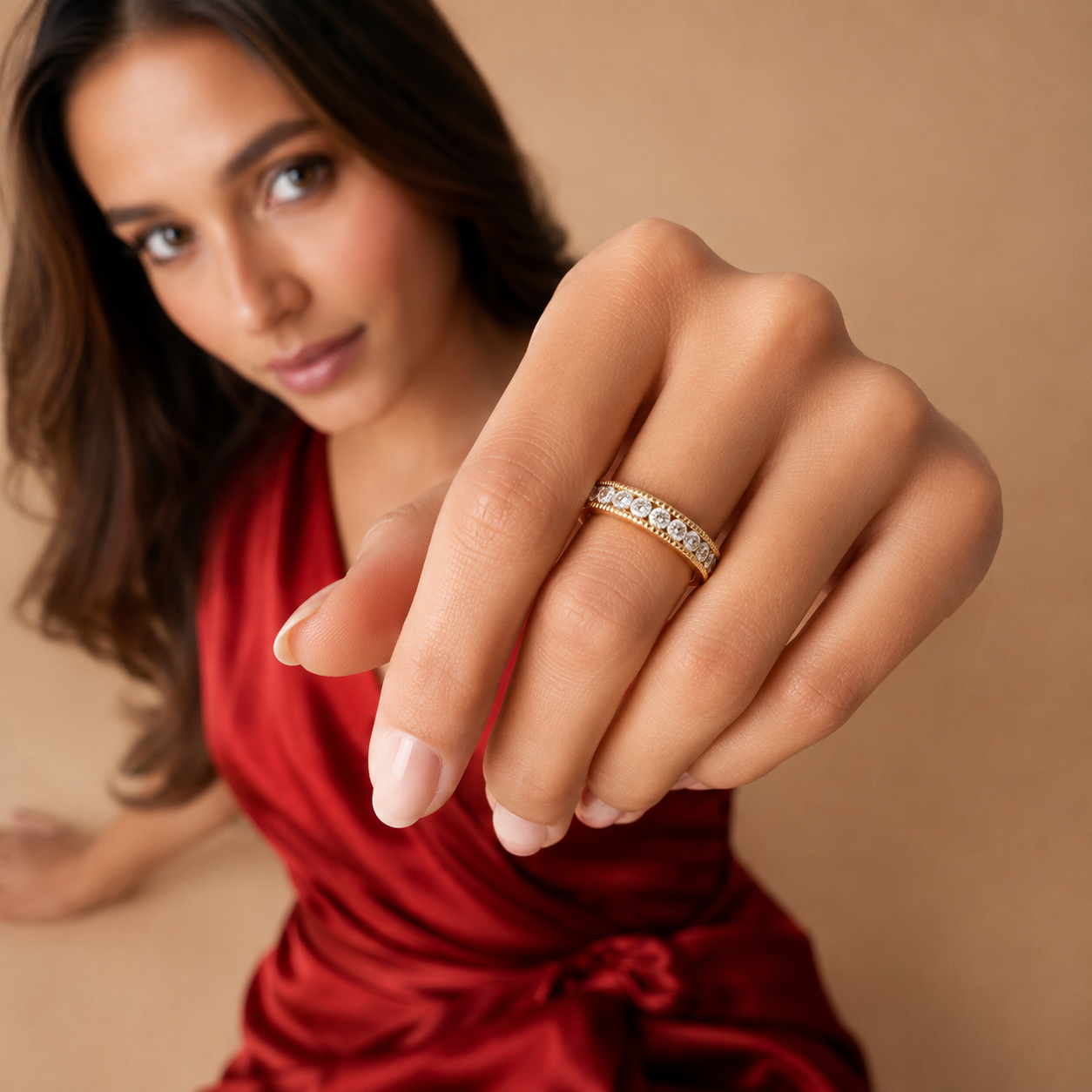 A close-up lifestyle shot of a woman wearing a thin gold band ring set with small clear stones, shown on her finger against a warm beige background.