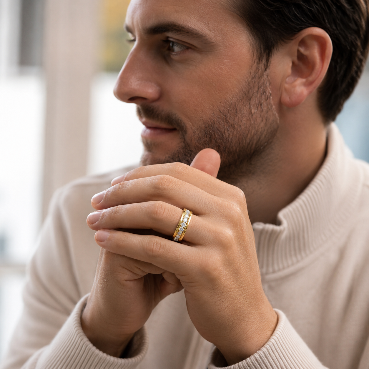 A close-up lifestyle shot of a man wearing a gold band ring with small clear stones on his ring finger, dressed in a cream-colored ribbed turtleneck sweater.