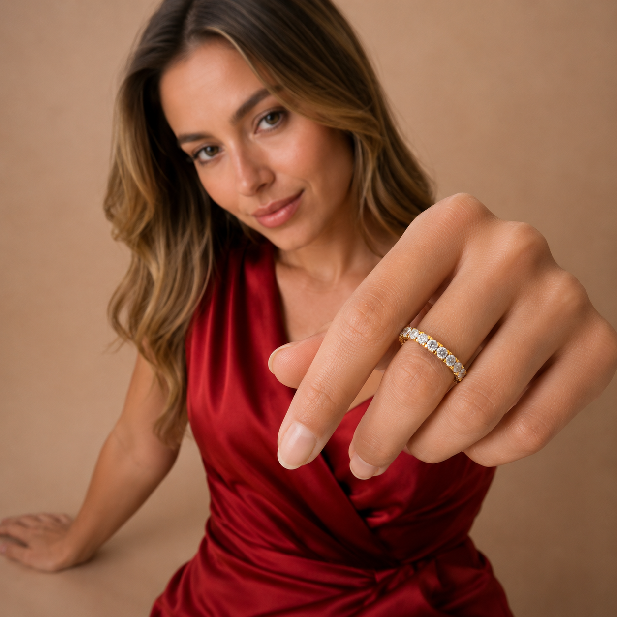 A woman in a red satin dress extends her hand to show a delicate silver ring set with small clear stones. The background is a solid, warm beige studio backdrop.